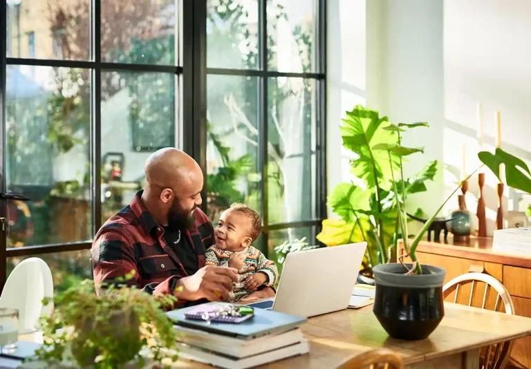 Smiling dad at his desk with his young daughter sitting on his lap, both looking and smiling at each other.