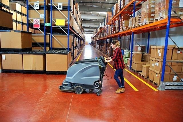 Person cleaning the floor in a warehouse with tall shelves and boxes stacked in the aisles.