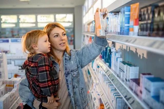 A woman holds a child in one arm while reaching for a product on a shelf in the pharmacy, her denim jacket catching the light. The child, dressed in a plaid shirt, looks on as she considers items amidst the bustling aisle filled with health and beauty products.