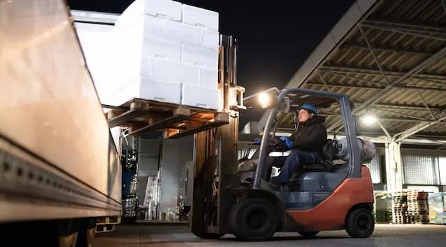A forklift truck loads stock in a warehouse at night.