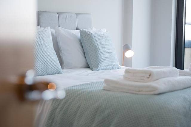 Neatly made bed in a cozy room with grey headboard, blue pillows, and white towels on the bed.