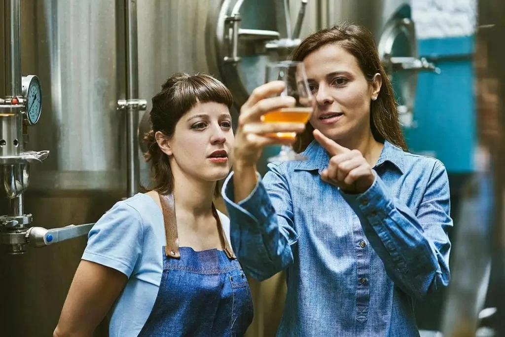 Two women stand in a brewery, discussing brewery insurance. One holds a glass of beer, observing its color while pointing at it with interest. Both wear blue clothing as large metal brewing tanks loom in the background.