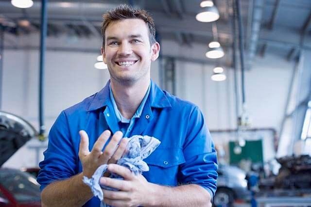 Mechanic in a blue uniform smiling and cleaning his hands with a cloth in a well-lit garage. Cars and mechanical equipment are visible in the background.