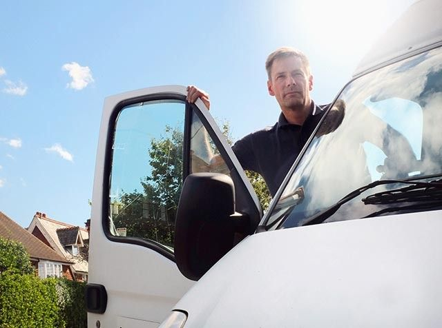 A person stands by the open drivers door of a white van under a clear blue sky. The sun is shining brightly, partially obscuring the persons face. Houses and greenery are visible in the background.